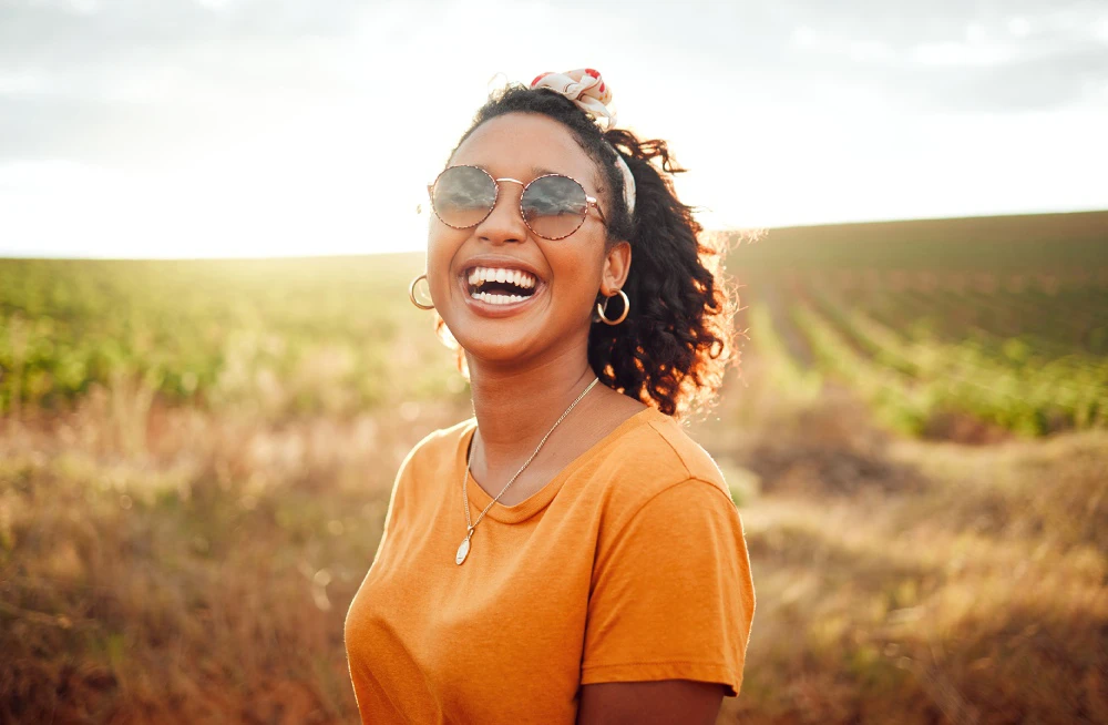Vibrant woman wearing sunglasses standing in front of a field with the sun radiating behind her